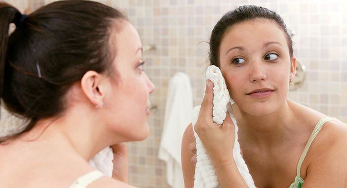 A young woman is looking at her reflection in a bathroom mirror while gently drying her face with a towel.