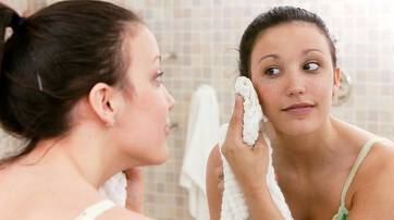 A young woman is looking at her reflection in a bathroom mirror while gently drying her face with a towel.