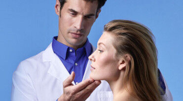 A healthcare professional examines the face of a patient in a clinical setting, with a light blue background.