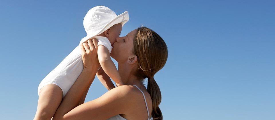 A woman holds a baby in the air and gives them a kiss against a clear blue sky.