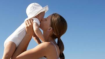A mother holding her baby high in the air, sharing a kiss against a clear blue sky.