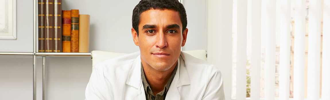 A young male doctor wearing a white lab coat, sitting at a desk in a well-lit office with bookshelves in the background.