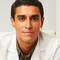 A young male doctor wearing a white lab coat, sitting at a desk in a well-lit office with bookshelves in the background.