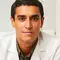 A young male doctor wearing a white lab coat, sitting at a desk in a well-lit office with bookshelves in the background.