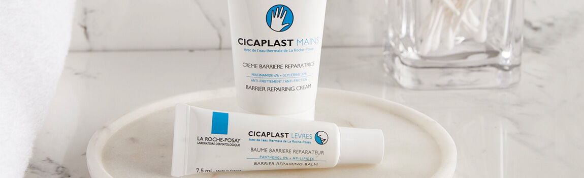 Two tubes of cream are displayed on a marble surface. One is labeled "Cicaplast Mains" and the other "Cicaplast Levres," both indicating they are barrier repairing products. A glass container with cotton swabs is visible in the background.