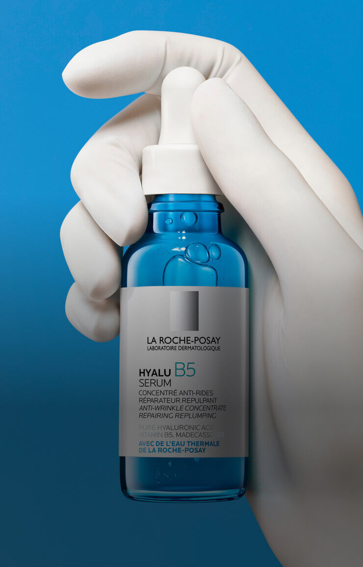 Flacon de serum Hyalu B5 de La Roche-Posay, tenu dans une main gantee, avec un fond bleu.