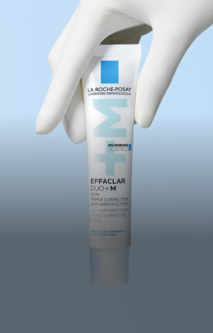 Un tube de creme Effaclar Duo+ M de La Roche-Posay, tenu par une main gantee, sur un fond bleu.