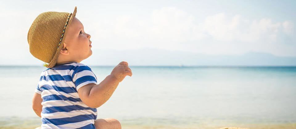 A baby wearing a straw hat and a striped shirt sits on the beach, looking up at the sky with the ocean in the background.