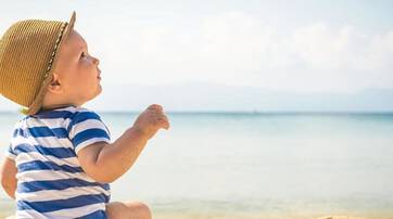 A baby wearing a straw hat and a striped shirt sits on the beach, looking up at the sky with the ocean in the background.
