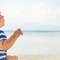A baby in a straw hat and a striped shirt sits on the beach, looking out at the calm ocean and distant mountains under a clear blue sky.