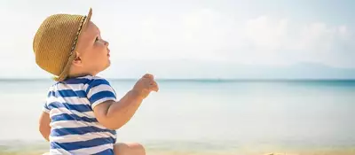 A baby in a straw hat and a striped shirt sits on the beach, looking out at the calm ocean and distant mountains under a clear blue sky.