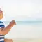 A baby in a straw hat and a striped shirt sits on the beach, looking out at the calm ocean and distant mountains under a clear blue sky.