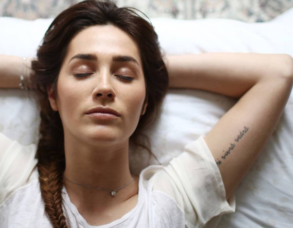 Une femme relaxee, les yeux fermes, repose sur un lit avec les bras derriere la tete.