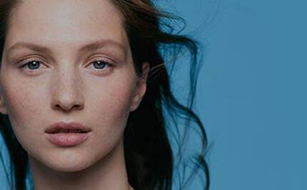 A close-up portrait of a person with long, flowing hair and a natural look, set against a plain blue background. They have a calm expression and visible freckles.