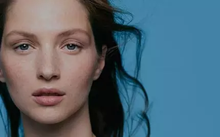 A close-up portrait of a person with long, flowing hair and a natural look, set against a plain blue background. They have a calm expression and visible freckles.