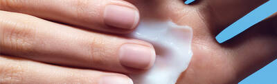 A close-up of a hand applying a creamy lotion to the palm, highlighting smooth, well-groomed nails against a light blue background.