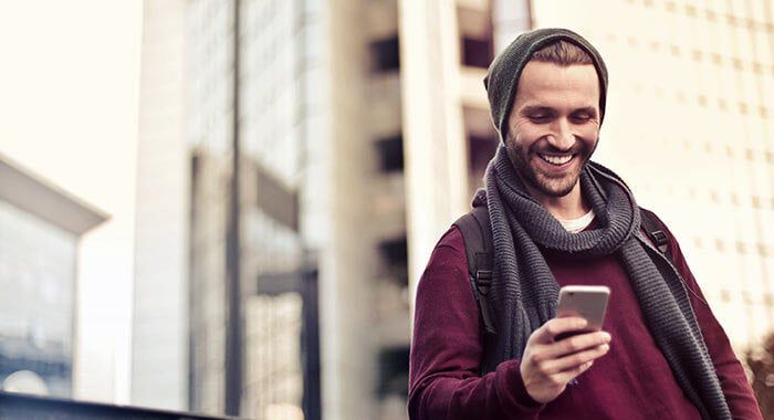 A smiling man wearing a beanie and scarf is standing outdoors, looking at his smartphone with modern buildings in the background.