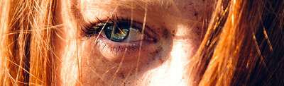 Close-up of a person's eye with striking blue irises and long eyelashes, framed by wavy red hair and scattered freckles on the skin.