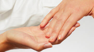 A close-up of two hands: one hand gently holds the fingertips of another, both appearing well-groomed against a soft, neutral background.