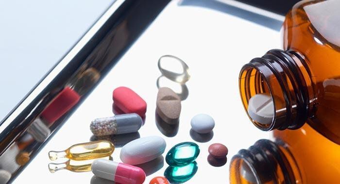 A variety of colorful pills and capsules are scattered around an open amber bottle on a reflective surface.