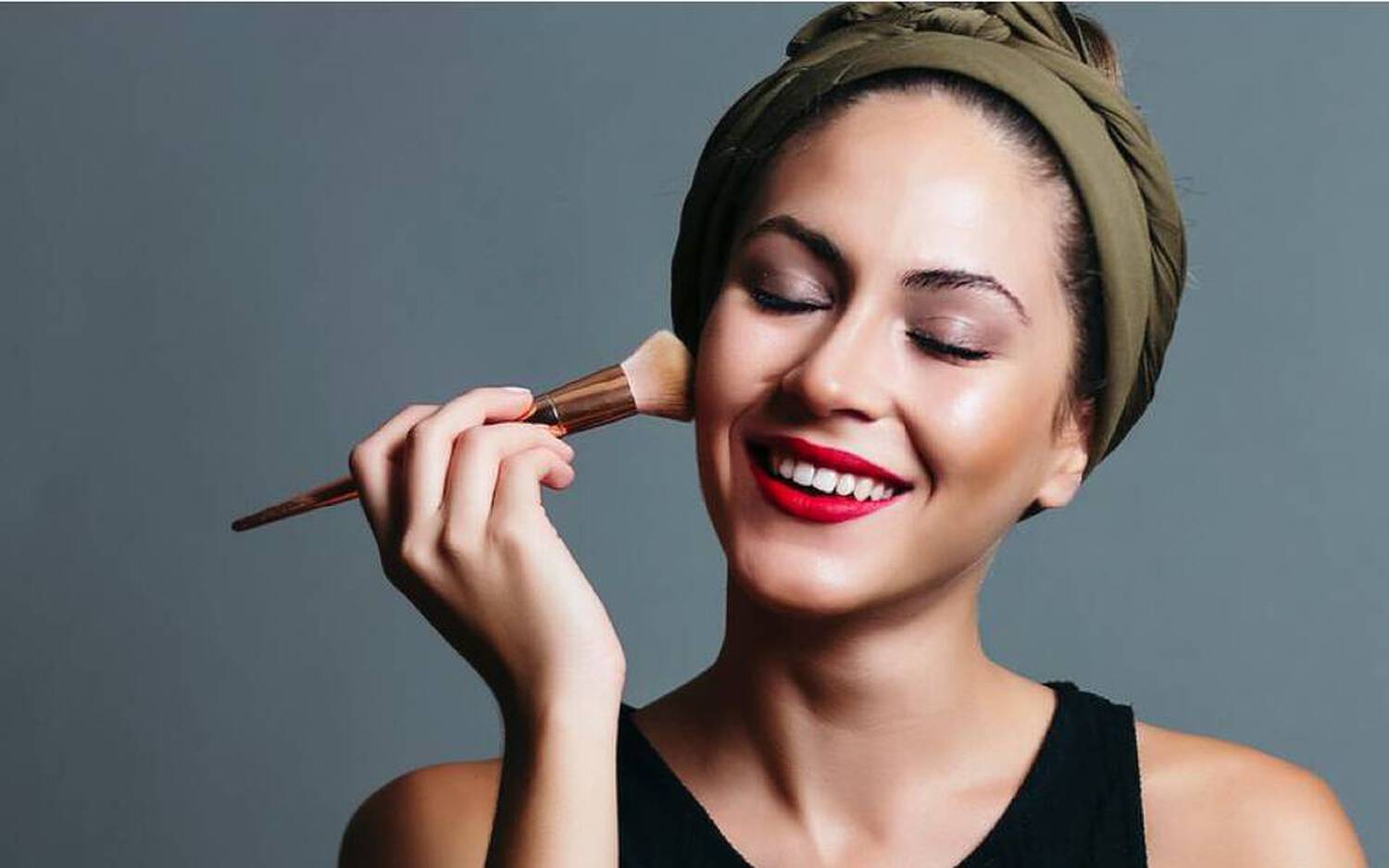 Femme souriante appliquant du maquillage sur son visage avec un pinceau, portant un bandeau vert.
