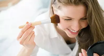 A woman smiling while applying blush with a makeup brush, looking into a handheld mirror.