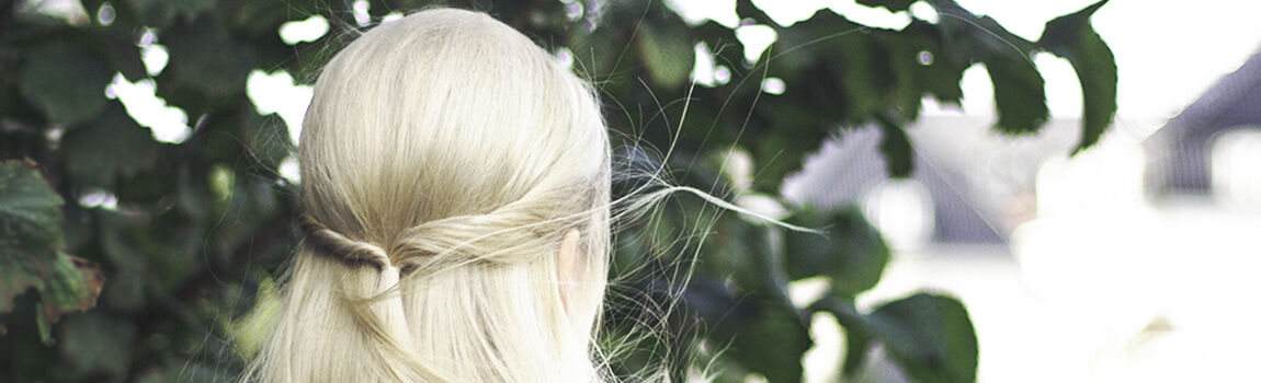 A person with long, light blonde hair styled in a half-up hairstyle, standing with their back to the camera in front of green foliage.