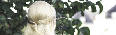 A person with long, light blonde hair styled in a half-up hairstyle, standing with their back to the camera in front of green foliage.