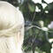 A person with long, light blonde hair styled in a half-up hairstyle, standing with their back to the camera in front of green foliage.