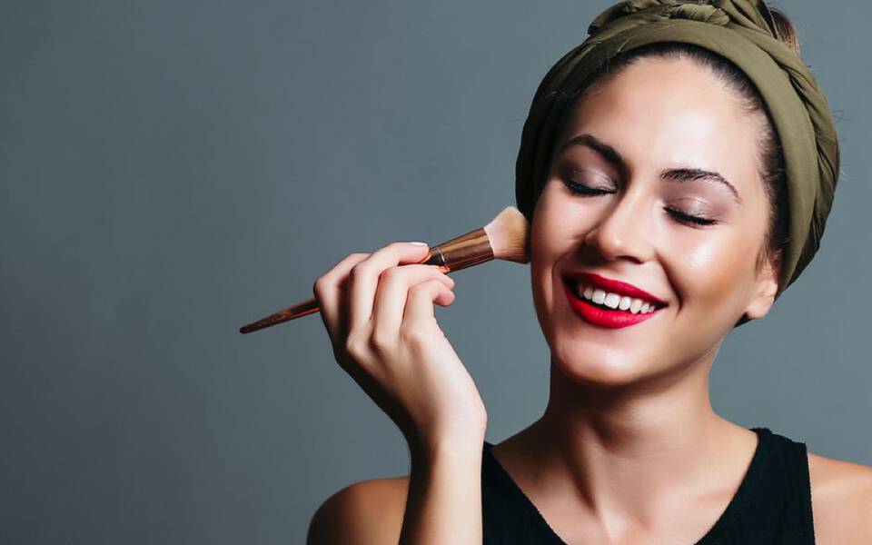 Une femme souriante applique du maquillage sur son visage avec un pinceau. Elle porte un bandeau et a des levres rouges.