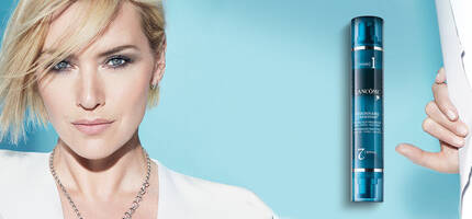 A close-up of a woman with short blonde hair and a striking expression, holding a skincare product against a turquoise background. The product is stylishly packaged with blue and silver tones.