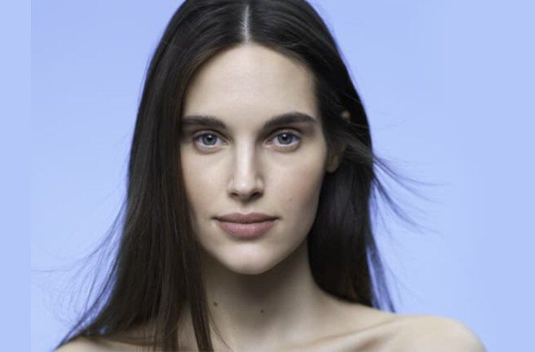 A close-up portrait of a woman with long, straight hair and a neutral expression, set against a soft blue background.