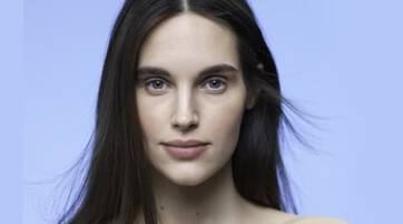 A close-up portrait of a woman with long, straight hair and a neutral expression, set against a soft blue background.