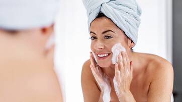 A woman in a towel wraps her hair while applying cleanser to her face in front of a mirror, smiling.