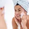 A woman in a bath towel applies facial cleanser on her face while looking in the mirror, smiling.