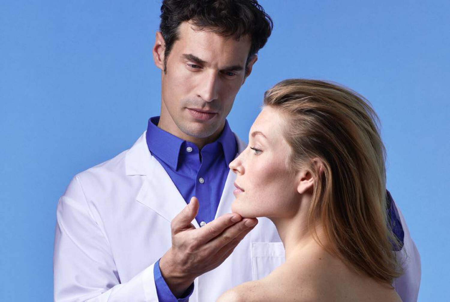 A healthcare professional examines a patient's face, with a blue background. The professional wears a white coat, and the patient has a relaxed expression.