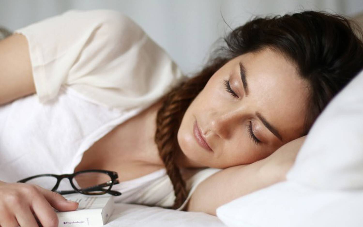 Une femme brune, allongee sur un lit, dort paisiblement avec des lunettes pres d'elle et une expression sereine.