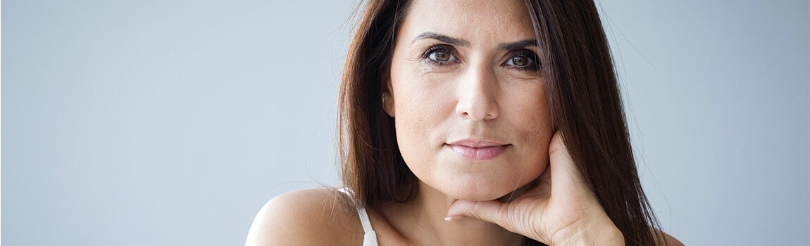 A close-up portrait of a woman with long, dark hair, resting her chin on her hand and looking thoughtfully at the camera against a light gray background.