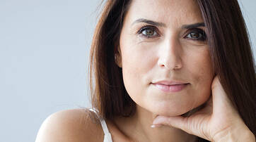 A close-up portrait of a woman with long, dark hair, resting her chin on her hand and looking thoughtfully at the camera against a light gray background.