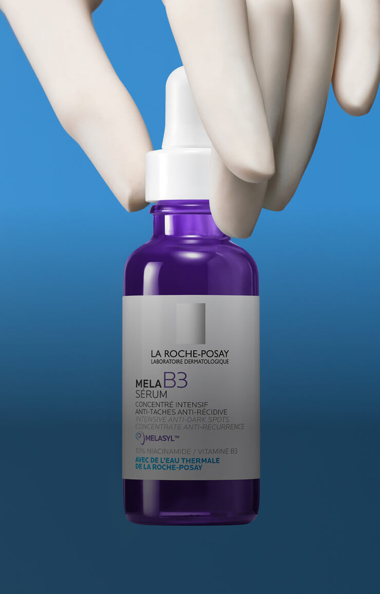 Flacon de serum MELA B3 de La Roche-Posay, tenant entre des doigts, sur fond bleu. Le serum est presente dans un flacon violet avec une etiquette en francais.