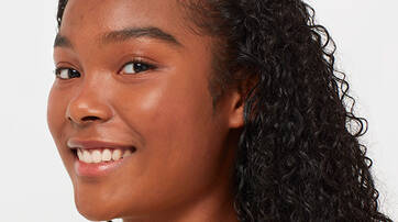 A young woman with curly hair smiles gently, facing slightly to the right against a neutral background.