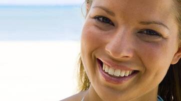 A smiling woman with long, light brown hair stands against a beach backdrop, with blue water and sand in the background.