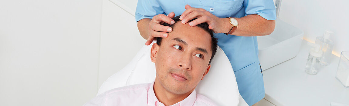 A man sits in a dental or medical chair while a healthcare professional gently examines his scalp. The setting is bright and clinical, with tools and supplies visible in the background.