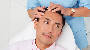 A person sitting in a treatment chair looks slightly concerned while another person gently examines their scalp. The atmosphere appears clinical and calming.