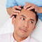 A man sits in a dental or medical chair while a healthcare professional gently examines his scalp. The setting is bright and clinical, with tools and supplies visible in the background.