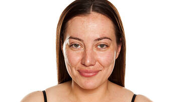 A young woman with long, straight brown hair smiles softly at the camera, wearing minimal makeup and a black top against a plain white background.