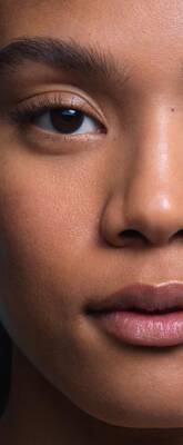 Une femme avec des cheveux frises, regardant l’appareil photo, a cote d'un smartphone affichant une image numerique de son visage.