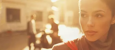 A close-up of a woman with a thoughtful expression, illuminated by warm sunlight, with blurred figures in the background.