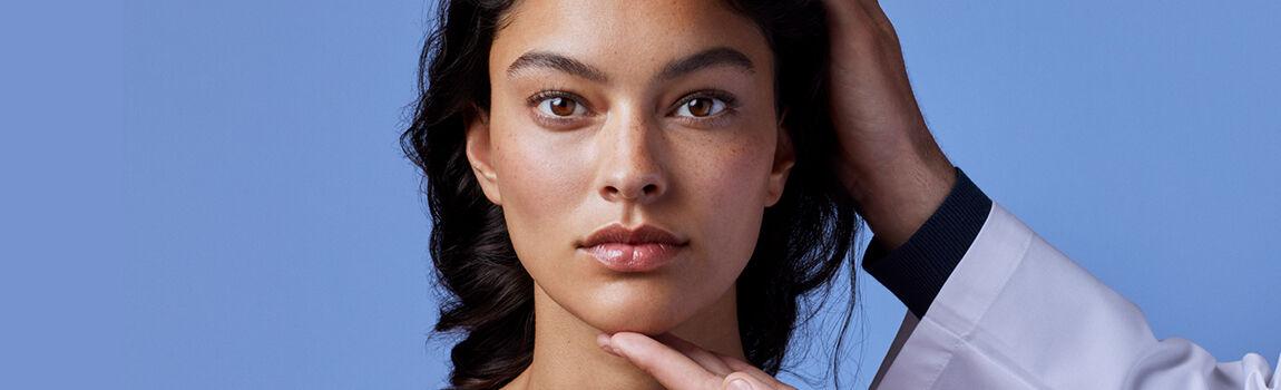 A close-up of a person with medium-toned skin and dark hair, looking directly at the camera. A hand, wearing a white coat, is gently touching their chin. The background is a soft blue.