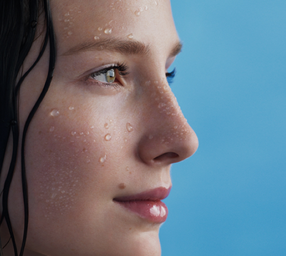 Portrait d'une femme avec des gouttes d'eau sur le visage, le regard tourne vers la droite, sur un fond bleu.